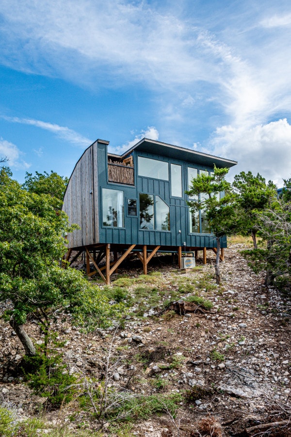 The exterior of the house displays a modern architectural design with large windows and a unique roofline. Set among native trees, the building is raised on stilts, providing an unobstructed view of the surrounding landscape. Natural stone and earth tones blend harmoniously with the environment.