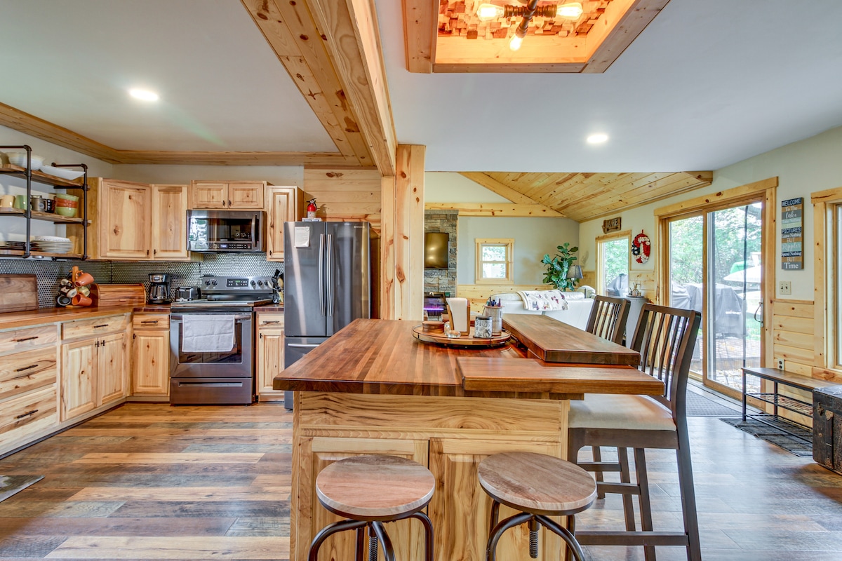 The kitchen area features wooden cabinetry and modern appliances, including a stainless steel refrigerator and oven. A spacious island with bar seating offers a gathering spot. Large windows allow natural light to fill the space, complemented by warm wood paneling on the ceiling.