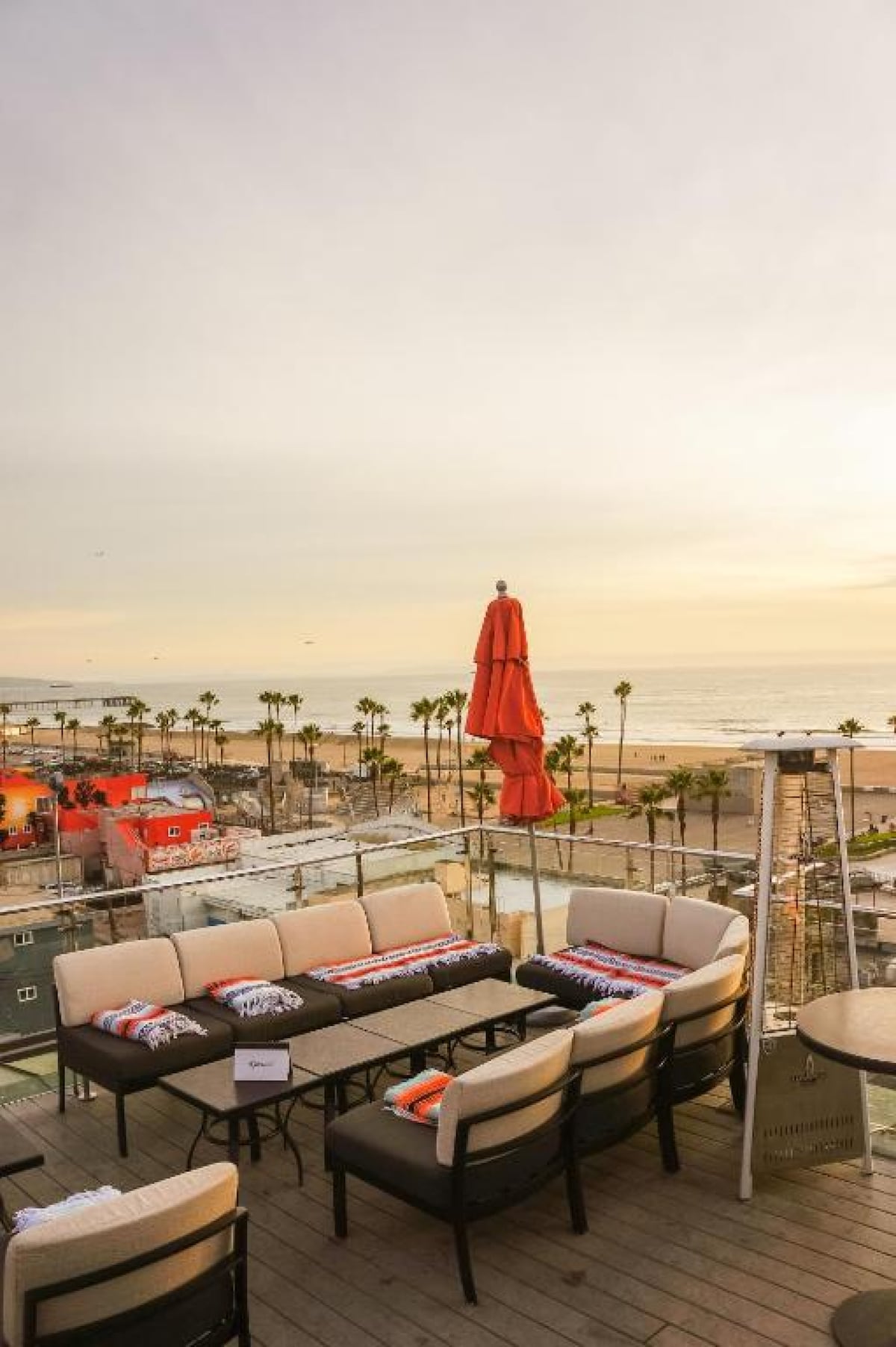 An open-air rooftop lounge is featured with stylish seating arranged around low tables. An umbrella provides shade, and palm trees can be seen in the background, along with a view of the beach and ocean under a soft evening sky.