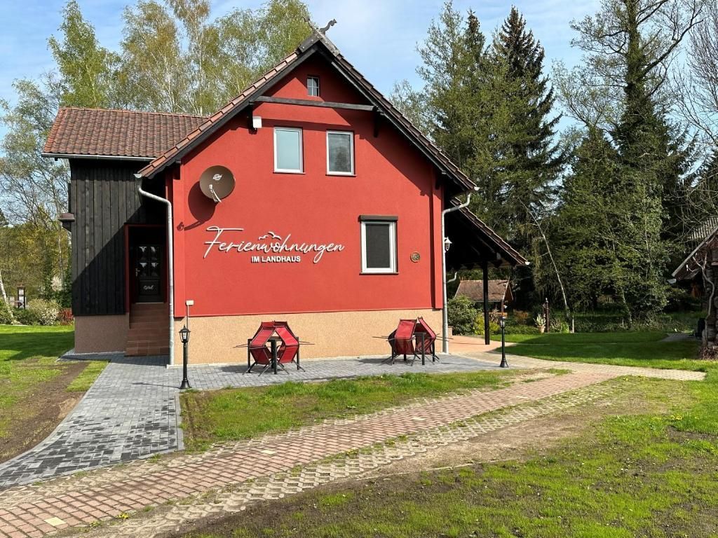 The exterior of a two-story building is presented, featuring a warm red facade with large windows. Seating areas with two red chairs and a small table are positioned on the front patio. Surrounding greenery includes trees and a grassy lawn, enhancing the tranquil outdoor space.