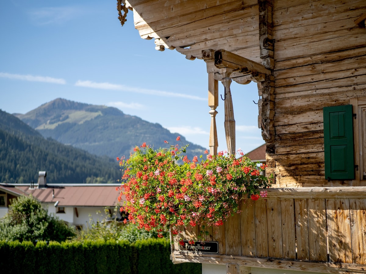 A wooden balcony adorned with vibrant flower boxes displays an array of colorful blooms. A backdrop of lush mountains under a clear blue sky enhances the rustic charm of the farmhouse.