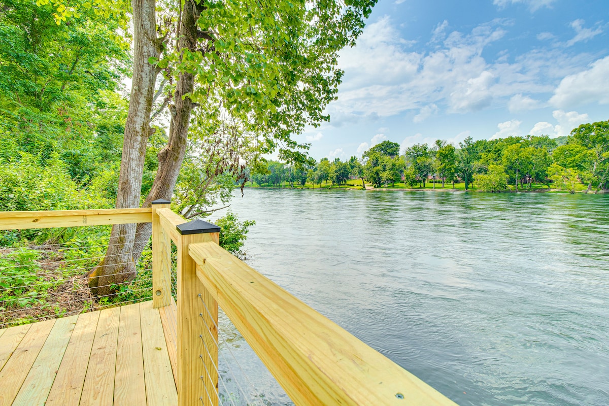 A wooden deck extends over the tranquil surface of the White River, surrounded by lush greenery and trees. The water reflects the blue sky and scattered clouds, creating a serene natural setting. The scene invites relaxation and appreciation of the riverside environment.