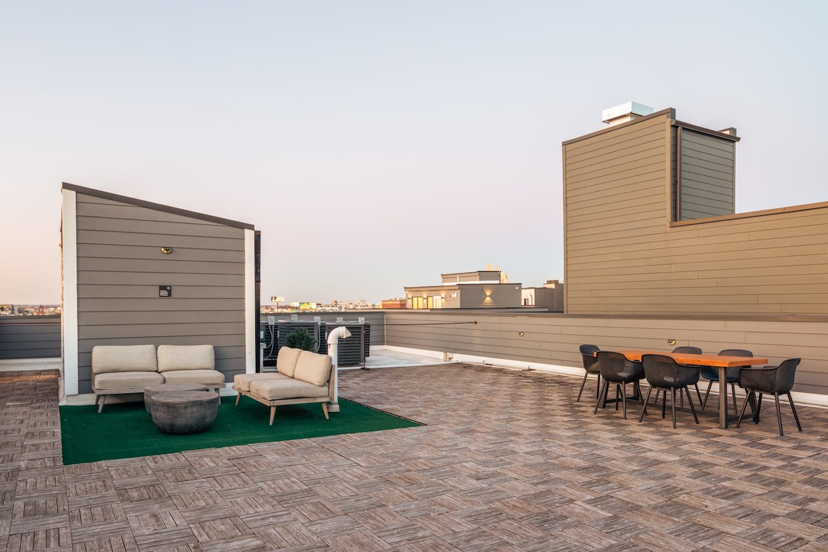 The shared roof-deck features stylish outdoor furniture on a smooth surface, with a seating area comprising a sofa and a round coffee table. A dining table and chairs are positioned nearby, complemented by views of the expansive skyline and surrounding rooftops.