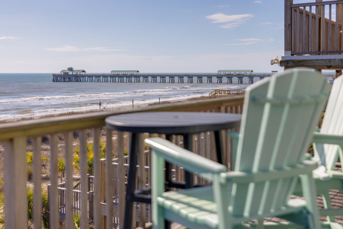 A view from a deck showcases the ocean and Folly Beach Pier, framed by a railing. Two mint-colored lounge chairs and a small black table are positioned in the foreground, inviting guests to enjoy the coastal scenery and breeze.