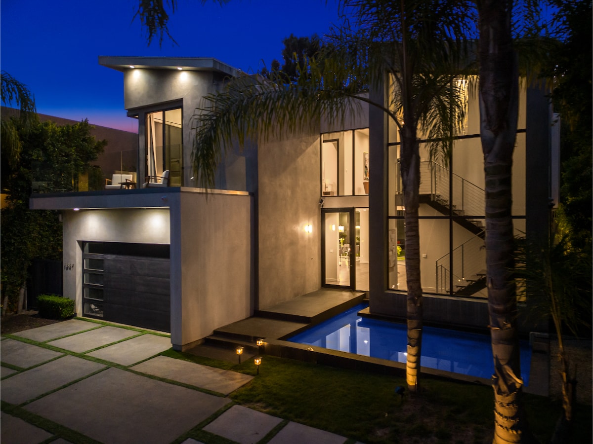 The exterior of a modern residence is illuminated at night, highlighting clean lines and expansive windows. A serene pool is visible beside the structure, reflecting the surrounding greenery, while stepping stones lead to the entrance, framed by elegant palm trees.