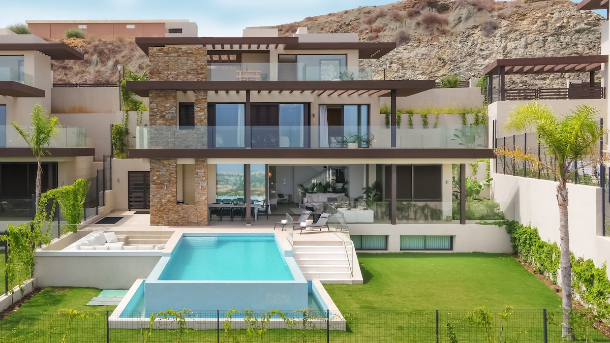 The villa’s exterior features a modern design with large glass windows, allowing natural light to fill the living spaces. A shimmering pool is situated on a sunlit terrace, surrounded by lush greenery and elegantly arranged landscaping.