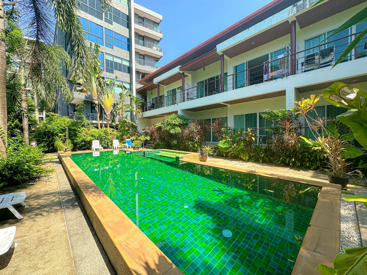 A swimming pool is surrounded by lush greenery and tropical plants, featuring a vibrant green tiled surface. Sun loungers are positioned nearby for relaxation, while a backdrop of multi-story buildings adds to the setting.