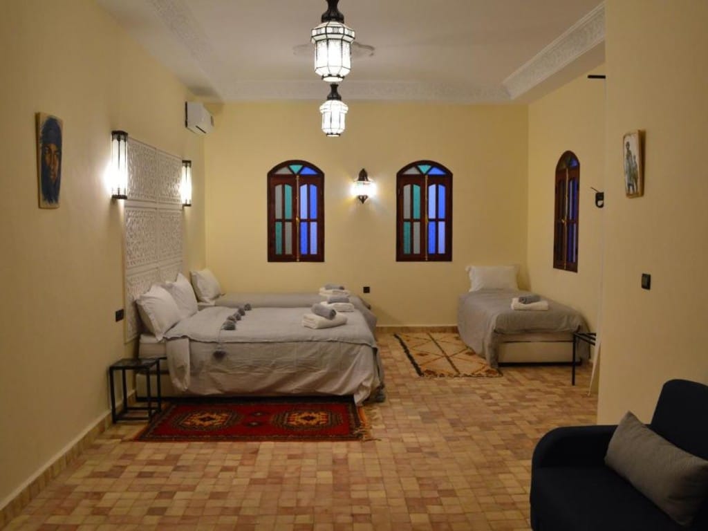A spacious guest room features two beds, one with a light duvet and the other with a neutral cover. Decorative wall art is displayed, and traditional lanterns provide soft lighting. Windows with decorative shutters allow natural light to enter, complementing the warm-colored walls.
