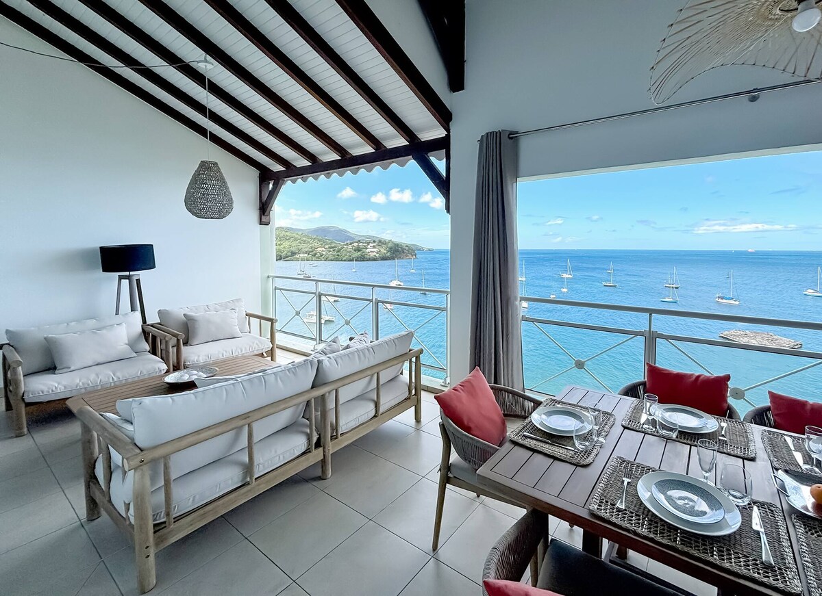 An open living area features comfortable seating options and a dining table with ocean views. Large windows allow natural light to fill the space, while the coastal scenery encompasses sailboats on the water and a distant shoreline, creating a serene environment.