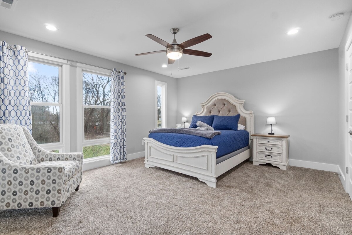 A spacious bedroom features a large bed with a blue comforter. Two windows allow natural light to enter, framed by patterned curtains. A minimalist nightstand and lamp complement the decor, while a textured chair adds a comfortable seating option.