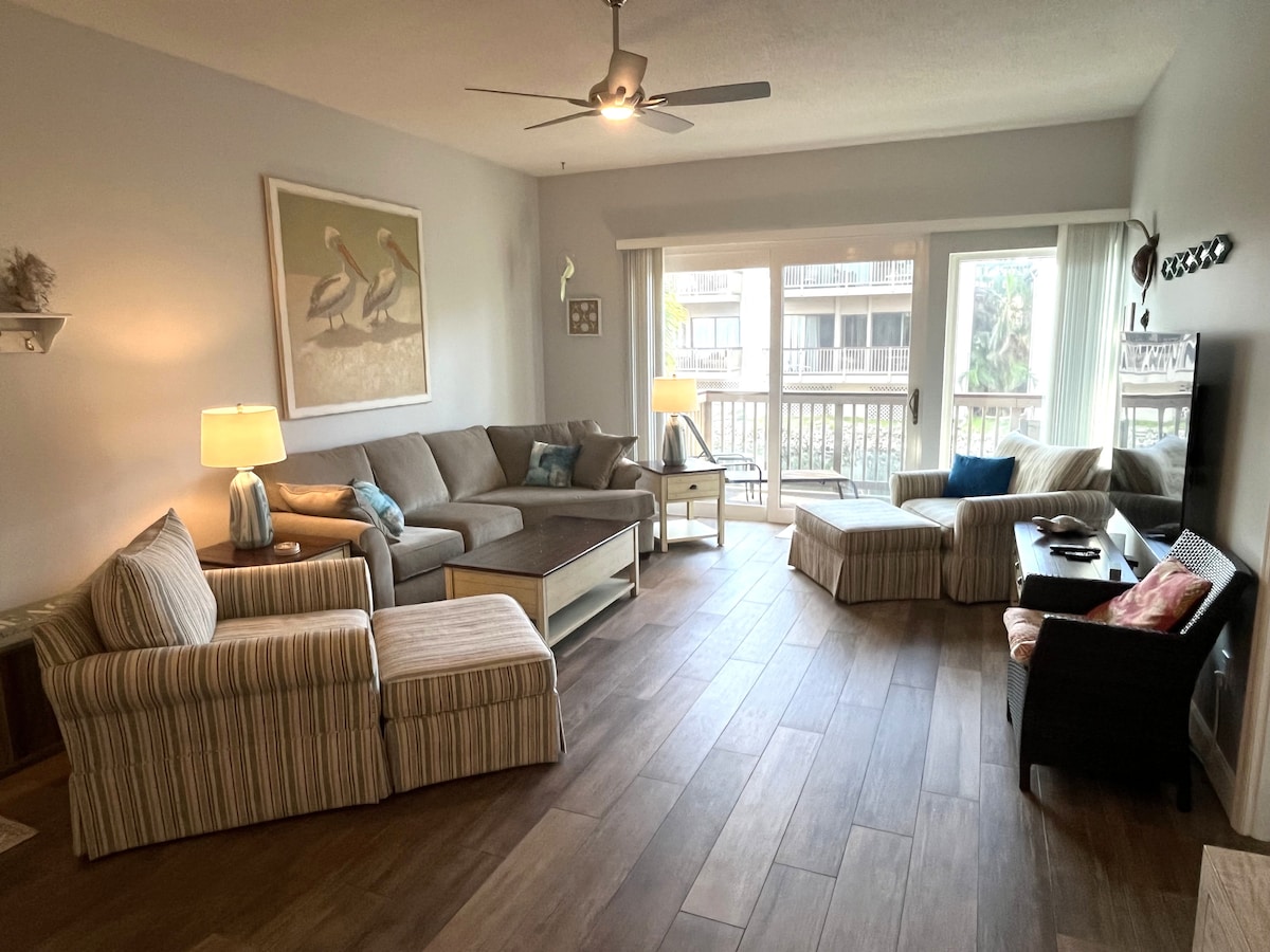 A spacious living room is highlighted by a large sectional sofa and additional seating, with natural light filtering through sliding glass doors. A decorative coastal artwork adorns the wall, while a ceiling fan enhances airflow. A flat-screen TV is positioned on a console table.