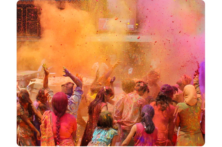 Festival Holi di India. Sekelompok besar orang, di tengah perayaan, terlihat dari belakang di tengah kabut warna-warni merah muda, oranye, dan ungu.