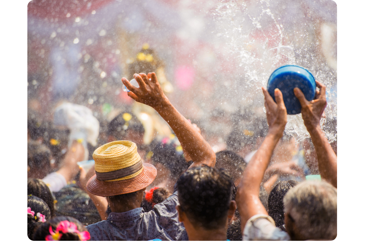 Festival Songkran di Thailand. Sekelompok besar orang yang membelakangi kita, mengangkat lengan mereka sambil memegang ember dan botol lalu menyiramkan air ke segala arah, membasahi semua orang.